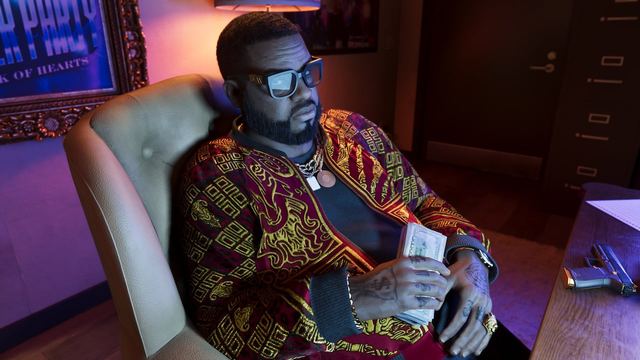 Boobie holding a stack of cash sitting back in a chair in front of a desk with a handgun on top of it.