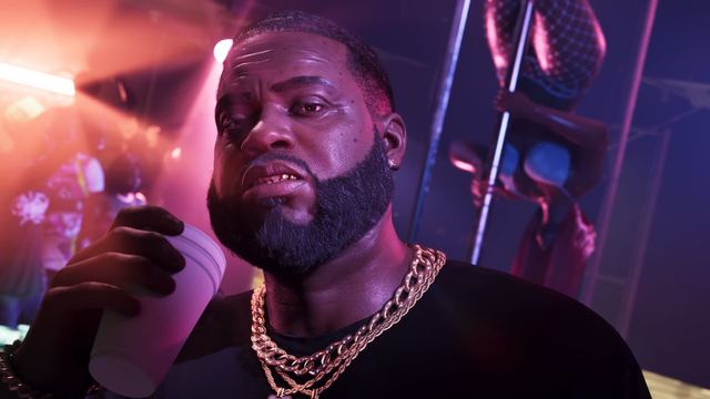 Boobie in a club holding a drink near his face. In the background a dancer is upside down on a pole.